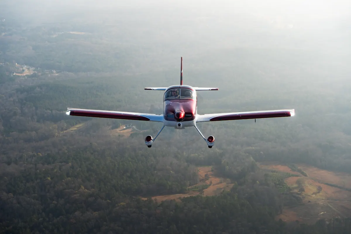 The red-and-white low-wing homebuilt flies straight at the camera, small against hazy forest and fields