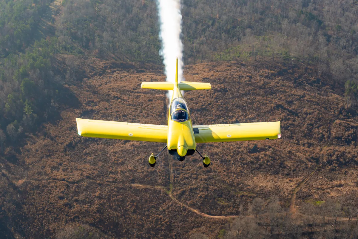 The yellow low-wing homebuilt flies toward the camera trailing white smoke, over cleared brown land