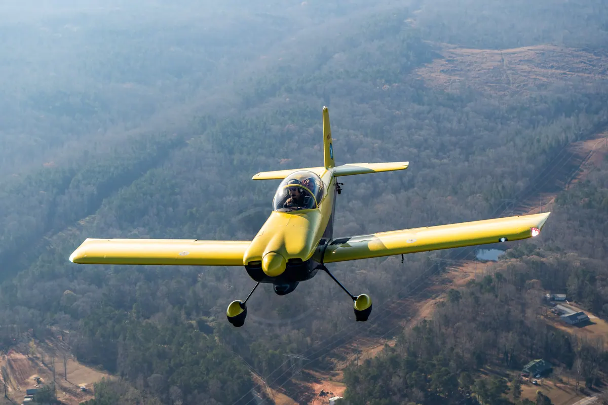 The yellow low-wing homebuilt banks toward the camera over hills and scattered houses