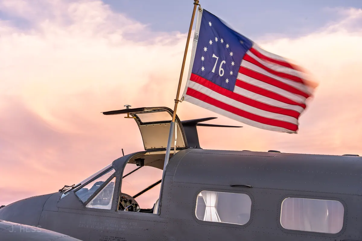 A Bicentennial '76' flag flies from the open cockpit hatch of an olive-drab Beech 18 against pastel sunset clouds