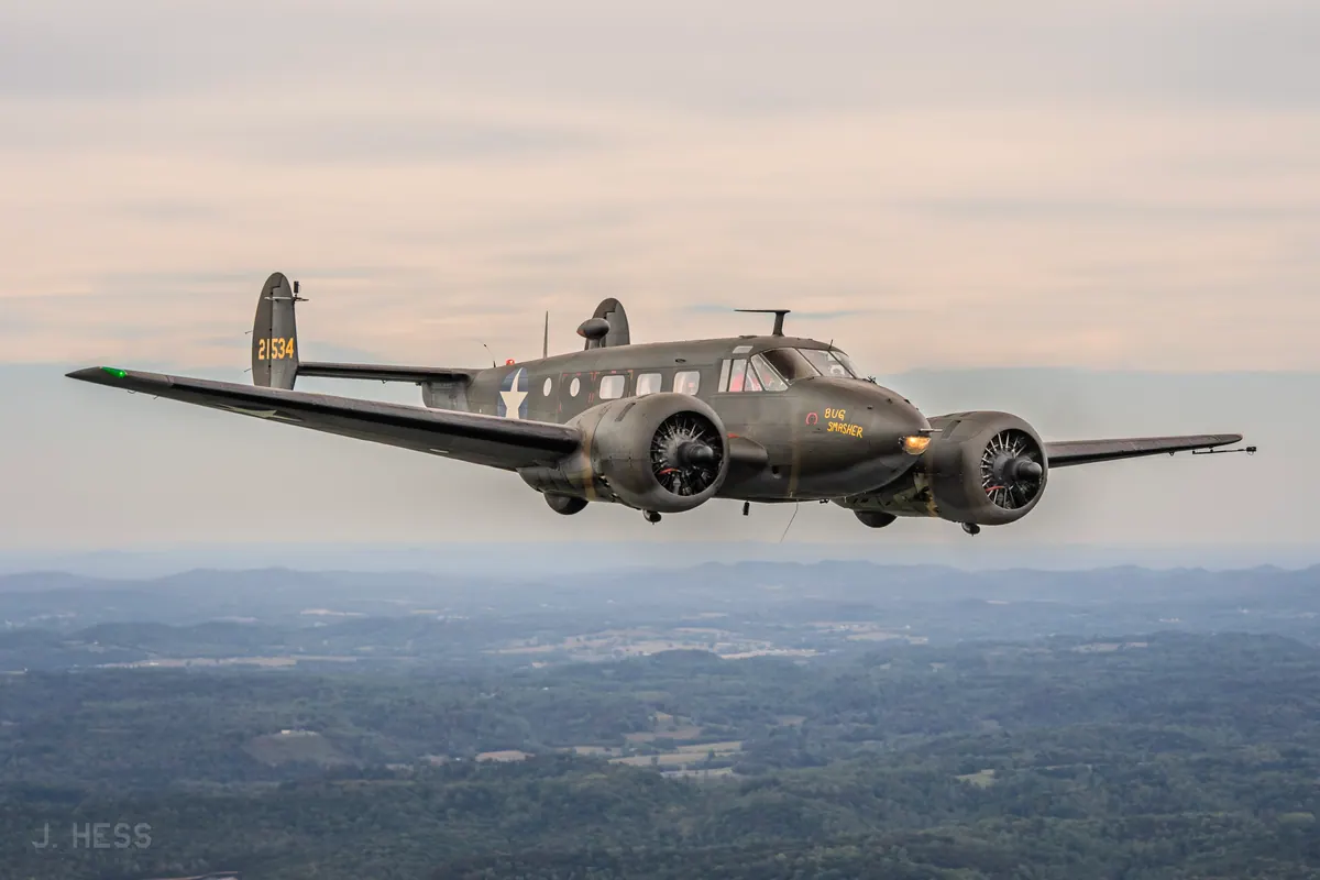 Side view of the olive-drab Beech 18 'Bug Smasher', serial 21534 on its tail and a star insignia on the fuselage, flying above hazy hills under a pale evening sky