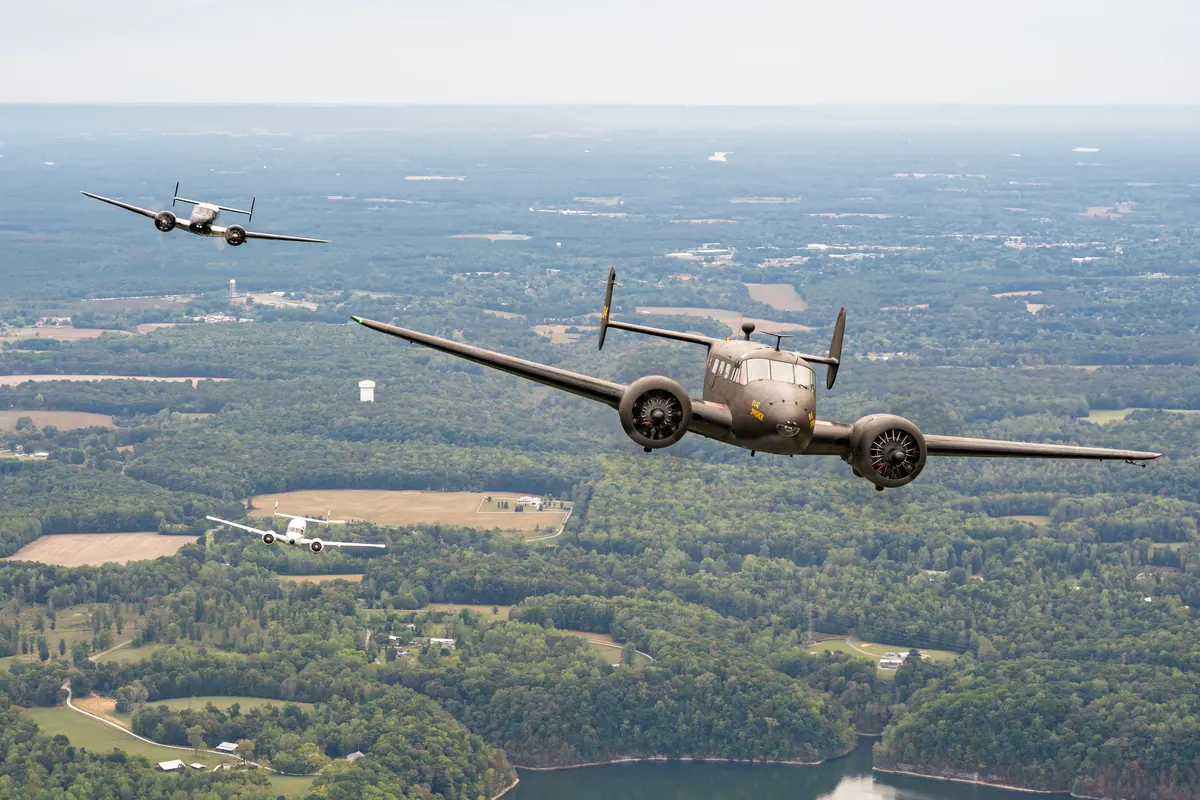 An olive-drab Beech 18 flies ahead of a silver and a white Twin Beech above wooded country and a lake