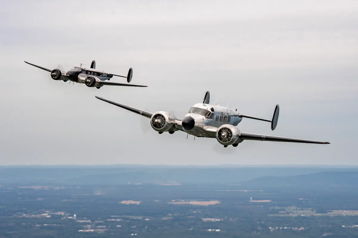 Two polished-aluminum Beech 18s flying in close formation against a pale gray sky above hazy farmland