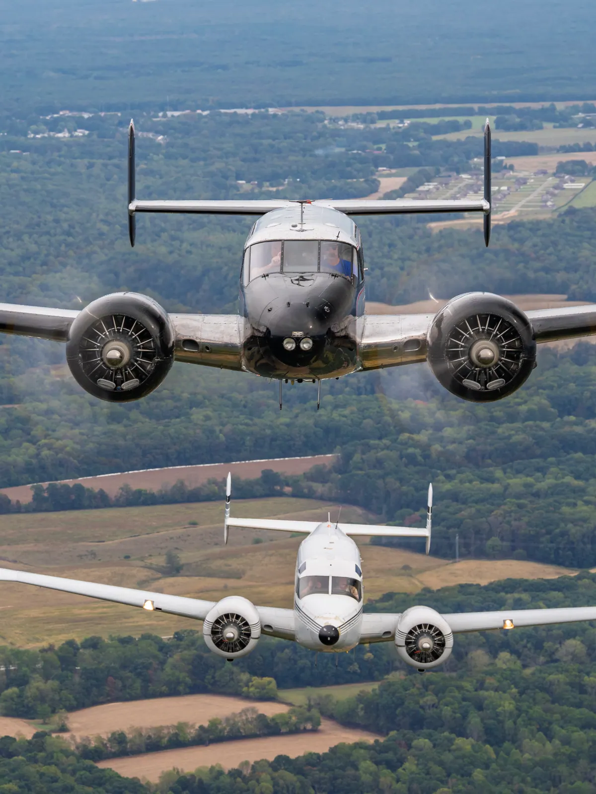A polished Beech 18 flies straight toward the camera with a white Twin Beech close below and behind it, over forest and farmland