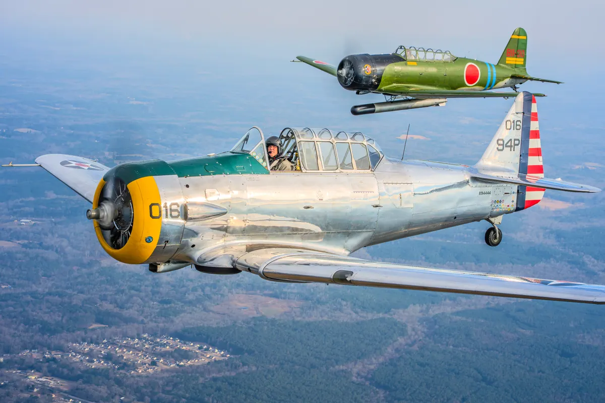The polished silver trainer '016' and the green 'Kate' torpedo-bomber replica fly together toward the camera over hazy blue woods