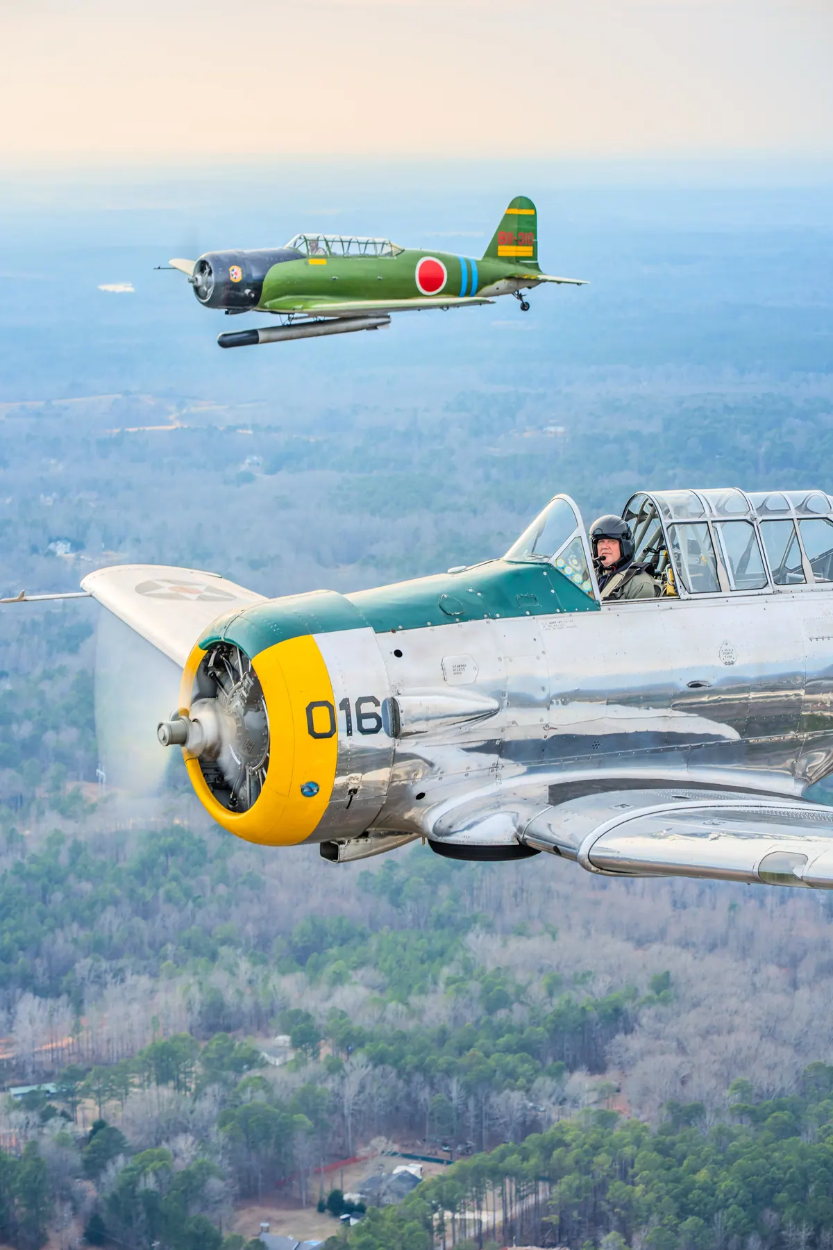 Vertical view: the polished silver trainer '016' in front, the green 'Kate' torpedo-bomber replica above, over wooded hills in warm late light